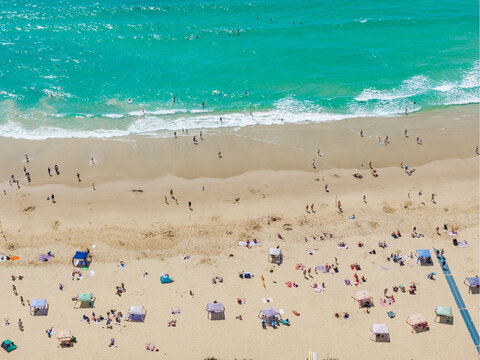 Aerial view of gentle waves on a white sandy beach covered in towels, umbrellas and sunshades