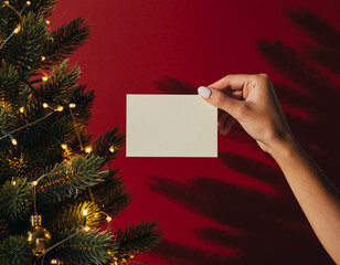 Tarjeta de felicitación navideña en blanco con mano femenina sobre fondo rojo junto a árbol decorado