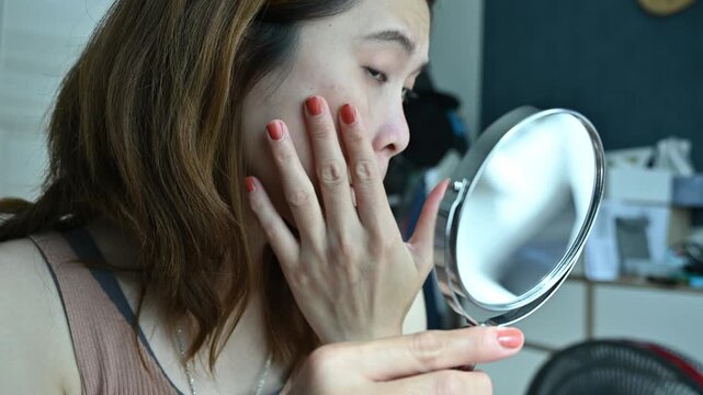 Asian woman using mirror to see acne and wrinkle problem on her face. Allergies, irritants, genetic makeup, etc can cause skin conditions.