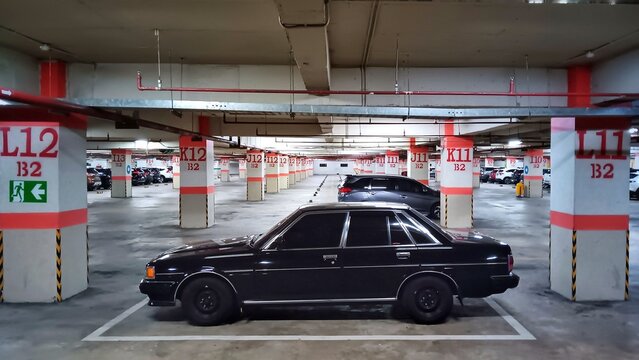 A wide-angle, low-light shot of a vintage black sedan parked in a deserted, multi-level concrete underground parking garage with prominent red and white numbered pillars, emphasizing the car's classic - Powered by Adobe