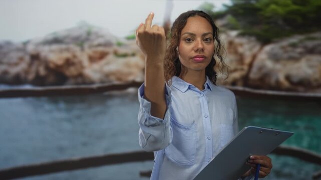 Young african american woman shows middle finger while holding clipboard and pen at rocky seaside railing; defiance protest boundary.