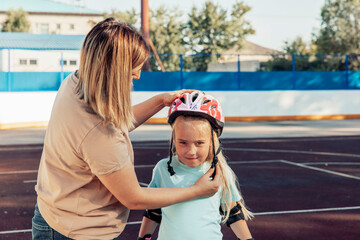 A woman helps a young girl put on a helmet at a skating rink. They are engaged in a bonding...
