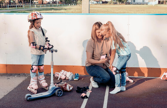 A mother assists her two young daughters during outdoor playtime. One girl is on a scooter, while the other is getting help with her roller skates.