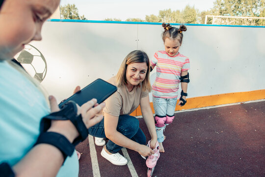 A mother helps her young daughter put on roller skates at a park. A young boy watches and records the moment with a smartphone. Family bonding and quality time are evident.