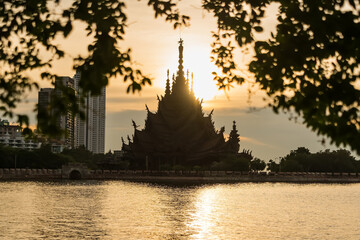 Chonburi, Thailand - November, 11, 2025 :Silhouette of an intricately carved wooden temple by the water at sunset framed by tree branches and modern buildings in the background