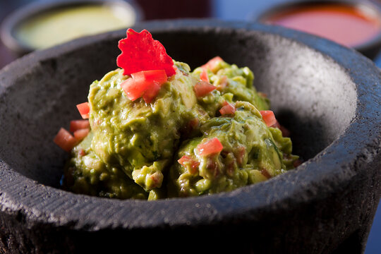 Chunky Mexican guacamole in a stone molcajete, accompanied by tomatoes and salsas in the background. 