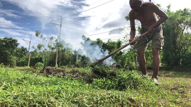 elderly farm worker is manually cultivating the soil with a hoe in the farmyard, despite thick white smoke from burning garbage.