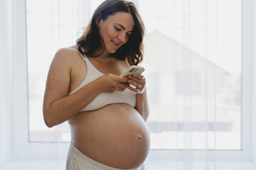 A pregnant woman stands by a bright window, smiling as she uses her phone. She is dressed in comfortable clothing, enjoying a serene moment in her home. This moment captures the beauty of pregnancy.