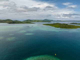 Lush green islands surrounded by shallow turquoise waters and coral patches under partly cloudy skies.