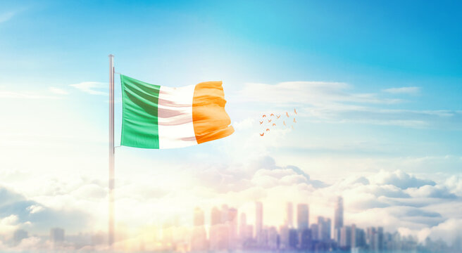 Ireland flag wavering in beautiful clouds with building skyline. The flag with drone shot in beautiful sky. Ireland national flag for independence day. - Powered by Adobe