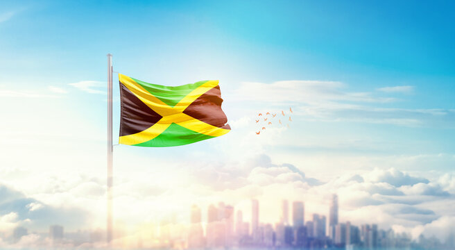 Jamaica flag wavering in beautiful clouds with building skyline. The flag with drone shot in beautiful sky. Jamaica national flag for independence day. - Powered by Adobe