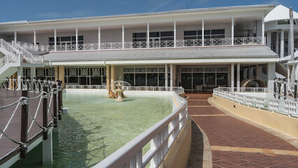 A tiled footpath with white railings runs along the turquoise water pool and fountain. The building is ahead. A boardwalk with rope railings nearby. Cuba. Varadero. Resort. Hotel.