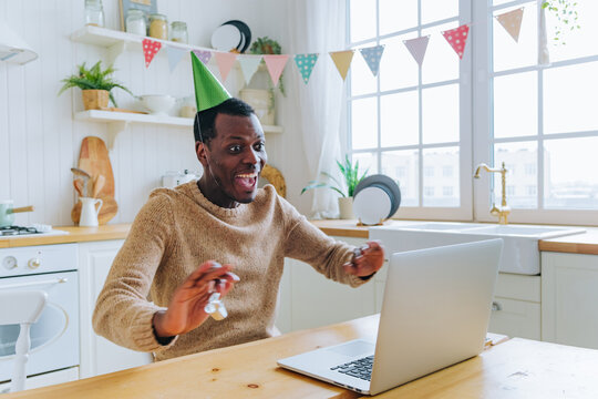 African american man happily celebrating his online birthday, smiling and making a joyful expression while interacting with friends and family on a laptop in a decorated kitchen