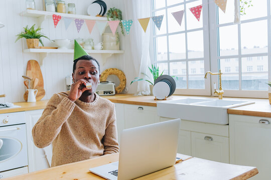 African american man in kitchen wearing a birthday hat, happily blowing a party horn during a virtual birthday video call on his laptop, connecting with loved ones remotely