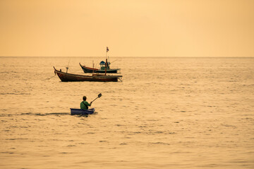 Chonburi, Thailand - November, 11, 2025 :Silhouetted fisherman paddling a small boat with traditional fishing boats anchored in the calm sea during golden hour