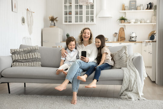 Joyful mum hugging two little kids on couch using phone