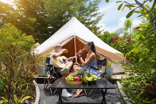 A man and woman is joyfully drinking beer and playing guitar near a cozy tent at glamping