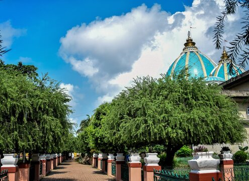 ISKCON the Indian Hindu Temple showcases its prominent blue and gold domes, set against a cloudy sky behind the green leaves and trees.