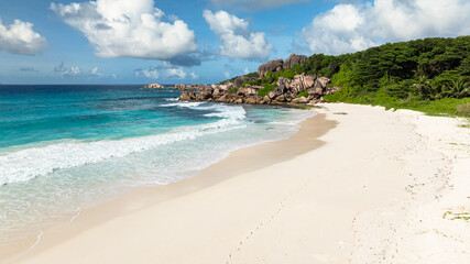 Waves washing the shore of a secluded beach with rocky cliffs and lush greenery. Grand Anse Beach, Seychelles..