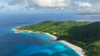 Turquoise ocean waves hitting a rocky shoreline bordered by green forested hills. La Digue, Seychelles.