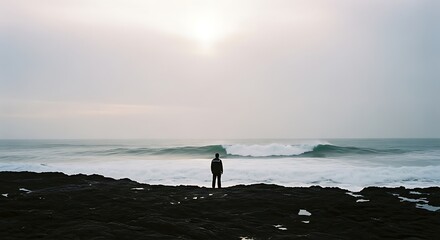 Lone figure stands on rocky shore watching ocean waves under overcast sky.