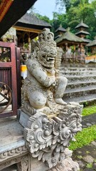 A stone statue of a deity sits at a temple entrance in Bali, Indonesia, with traditional architecture visible in the background.