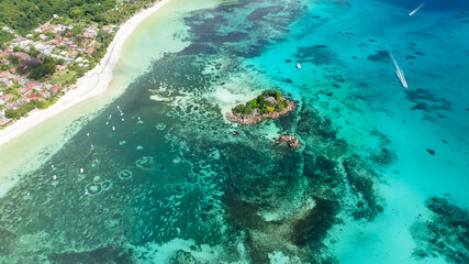 A small, lush island sits in shallow turquoise waters, surrounded by coral reefs and close to a white sandy shore. Praslin, Seychelles. © Alex Traveler