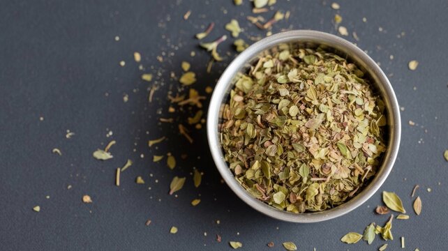 A small silver bowl brimming with dried oregano flakes, resting on a dark gray surface, with scattered herbs around.