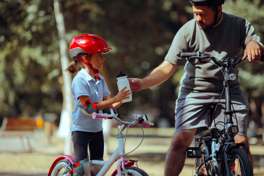 Dad Gives Little Cyclist a Water Bottle to Hydrate. Man helping his daughter to learn how to bike and stay hydrated 
