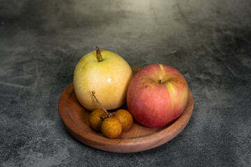 A pear, apple, and longan fruits are arranged in a wooden bowl on a textured gray surface, creating a simple natural still-life composition with warm tones and a clean rustic aesthetic.