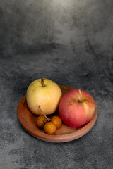 A pear, apple, and longan fruits are arranged in a wooden bowl on a textured gray surface, creating a simple natural still-life composition with warm tones and a clean rustic aesthetic.