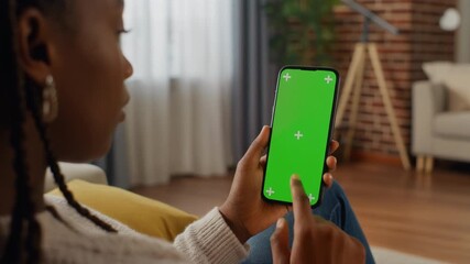 Over-the-shoulder shot of a young woman relaxing in a cozy apartment, swiping and tapping a vertical smartphone with a bright green screen (chroma key). Perfect for mockups.