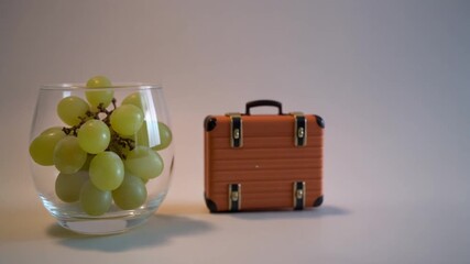 A glass bowl filled with fresh green grapes next to a miniature brown suitcase symbolizing a healthy snack for travel.