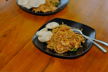 Overhead view of a serving of Indonesian Mie Goreng (fried noodles) with white crackers on a black plate, placed on a warm wooden table.