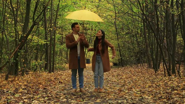 Smiling couple walking together under yellow umbrella in autumn park, wearing brown coats and jeans, enjoying romantic stroll among colorful fall trees and leaves.