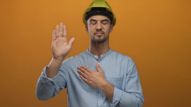 Young man wearing a hardhat making a pledge with one hand on chest and the other raised against an orange background, symbolizing commitment and honesty.
