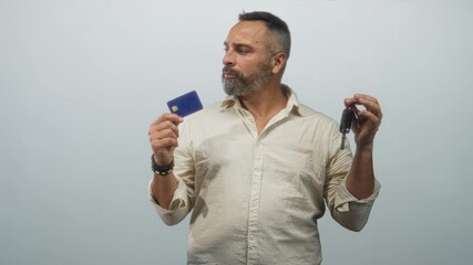 Man holding a creditcard and car keys in studio, comparing the two items while looking between them; choice doubt.
