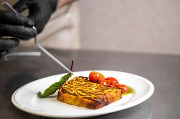 Chef plating gourmet layered dish with cherry tomatoes and green sauce. Professional food styling, fine dining presentation, and culinary expertise in a restaurant kitchen setting