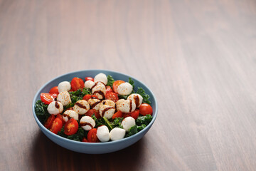 Blue bowl of salad with mozzarella, cherry tomatoes and kale, seasoned with balsamic sauce on walnut wood table