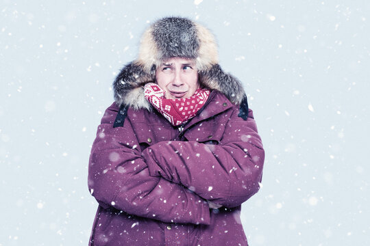 Man in red winter jacket and fur trapper hat shivers in falling snow, hugging himself for warmth against a cold, pale backdrop. Seasonal cold weather, freezing outdoor concept, harsh winter