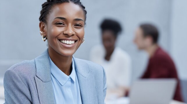 A confident woman with braided hair smiles warmly at the camera in a stylish suit. In the background, blurred colleagues engage in lively conversation, reflecting teamwork and collaboration