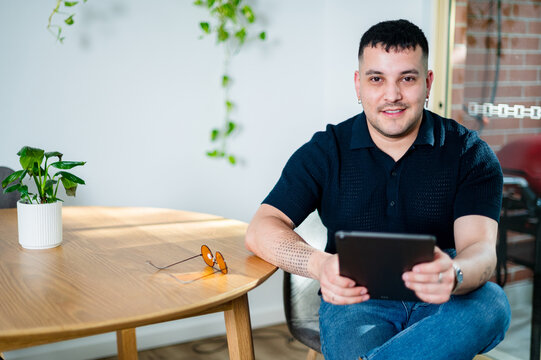 A young man sits comfortably at a wooden table, reading on a tablet device in his modern home.