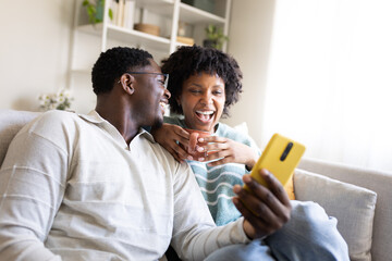 Happy couple watching funny videos on smartphone and laughing together on sofa at home