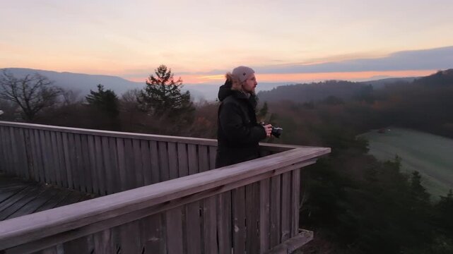 A serene morning scene high above the Swiss countryside. A lone photographer stands on a wooden lookout tower, taking pictures of the breathtaking landscape as the sun rises over mountains and valleys