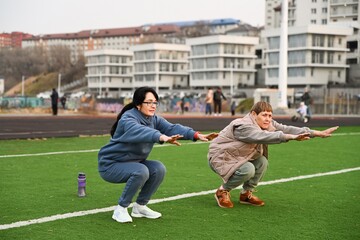 Two Russian women, aged 50 and 60, in tracksuits,
doing squats at the stadium
