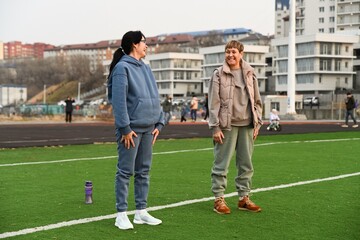 Two Russian women, aged 50 and 60, in tracksuits,
doing squats at the stadium and laughing