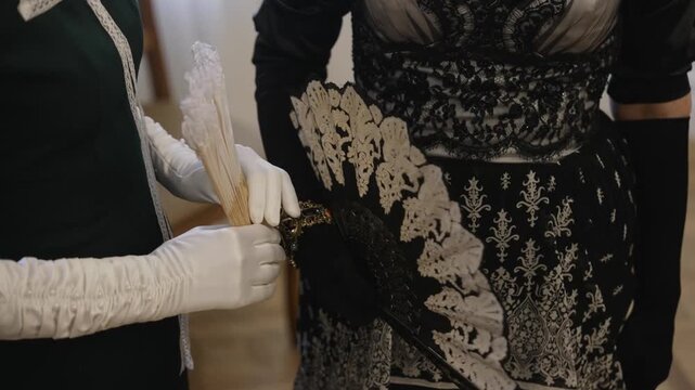 Two women wearing elegant historical dresses and white gloves holding vintage lace hand fans. Historical reconstruction of victorian era fashion and accessories
