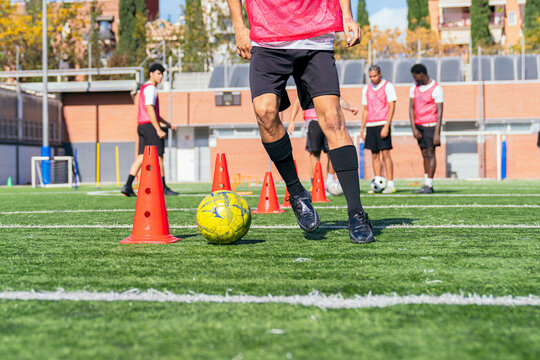 Football player dribbling soccer ball around training cones on a green field. Teammates practicing in background