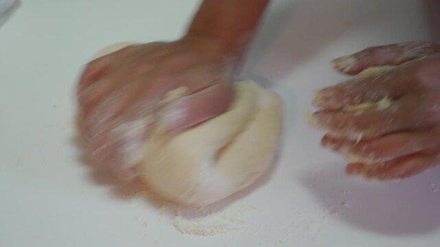 Hands covered in flour are vigorously kneading and pushing a smooth mass of light dough on a clean white countertop. The motion is dynamic and energetic, signifying final stage of dough preparation.