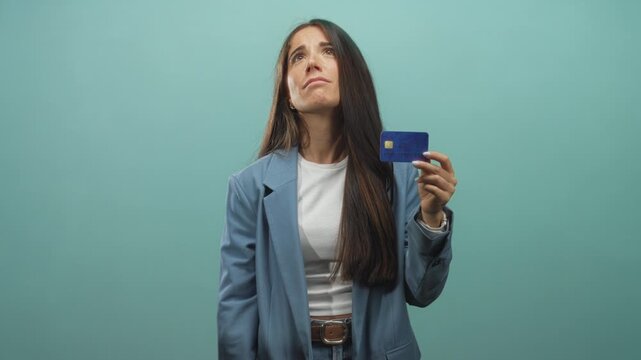 Woman holding blue creditcard up with concerned expression, presenting the card in studio wearing blue blazer and white top; financial anxiety.